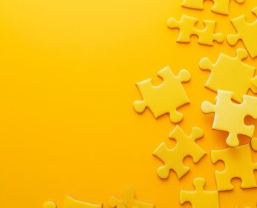An overhead view of multiple yellow puzzle pieces scattered haphazardly on a uniform yellow background, with ample copy space on the right for text.