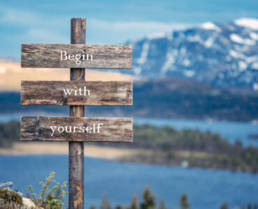 begin with yourself text quote written on wooden signpost outdoors in nature with lake and mountain scenery in the background. Moody feeling.